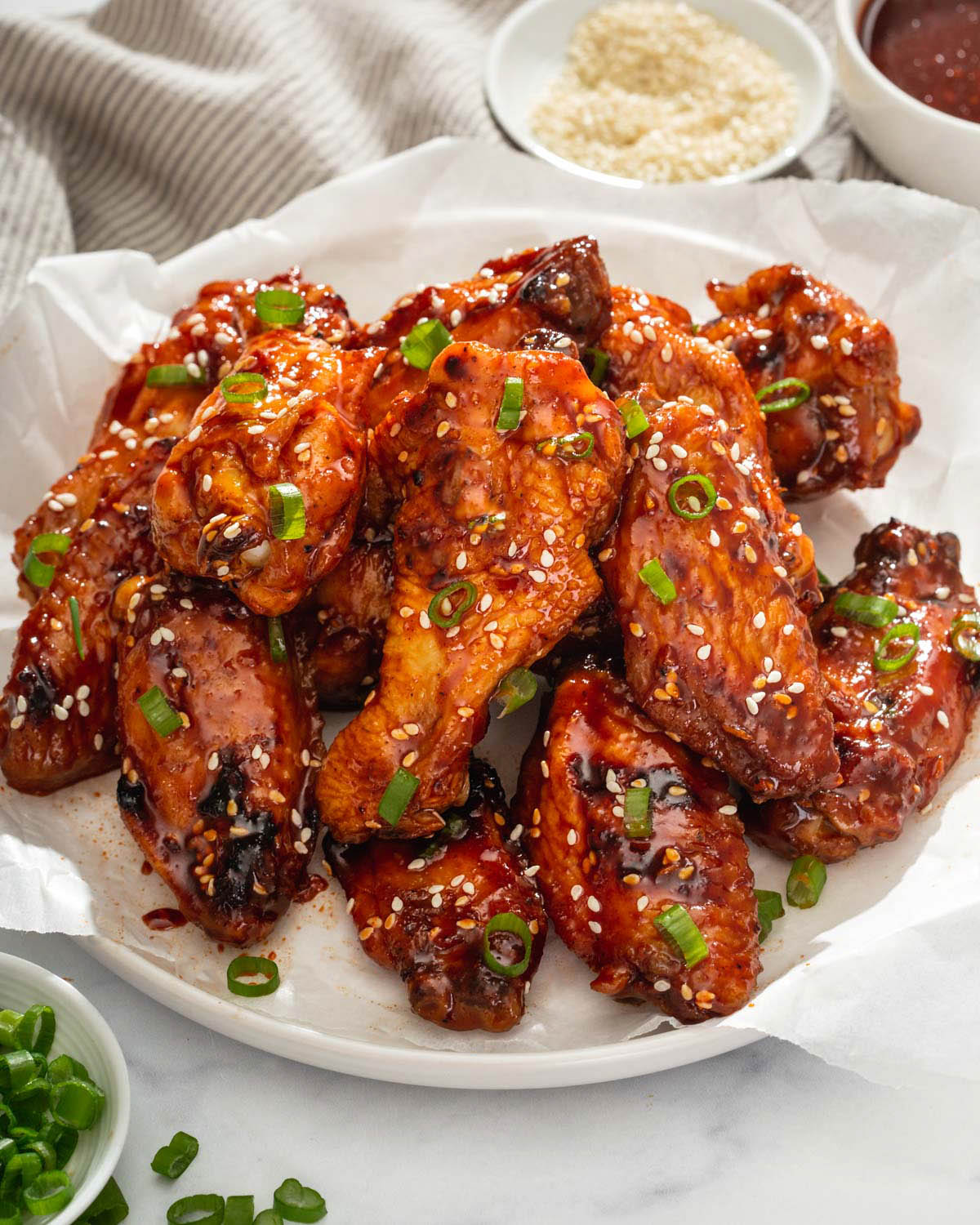 Close up of a plate of chicken wings surrounded by a linen, sauce, sesame seeds, and scallions.