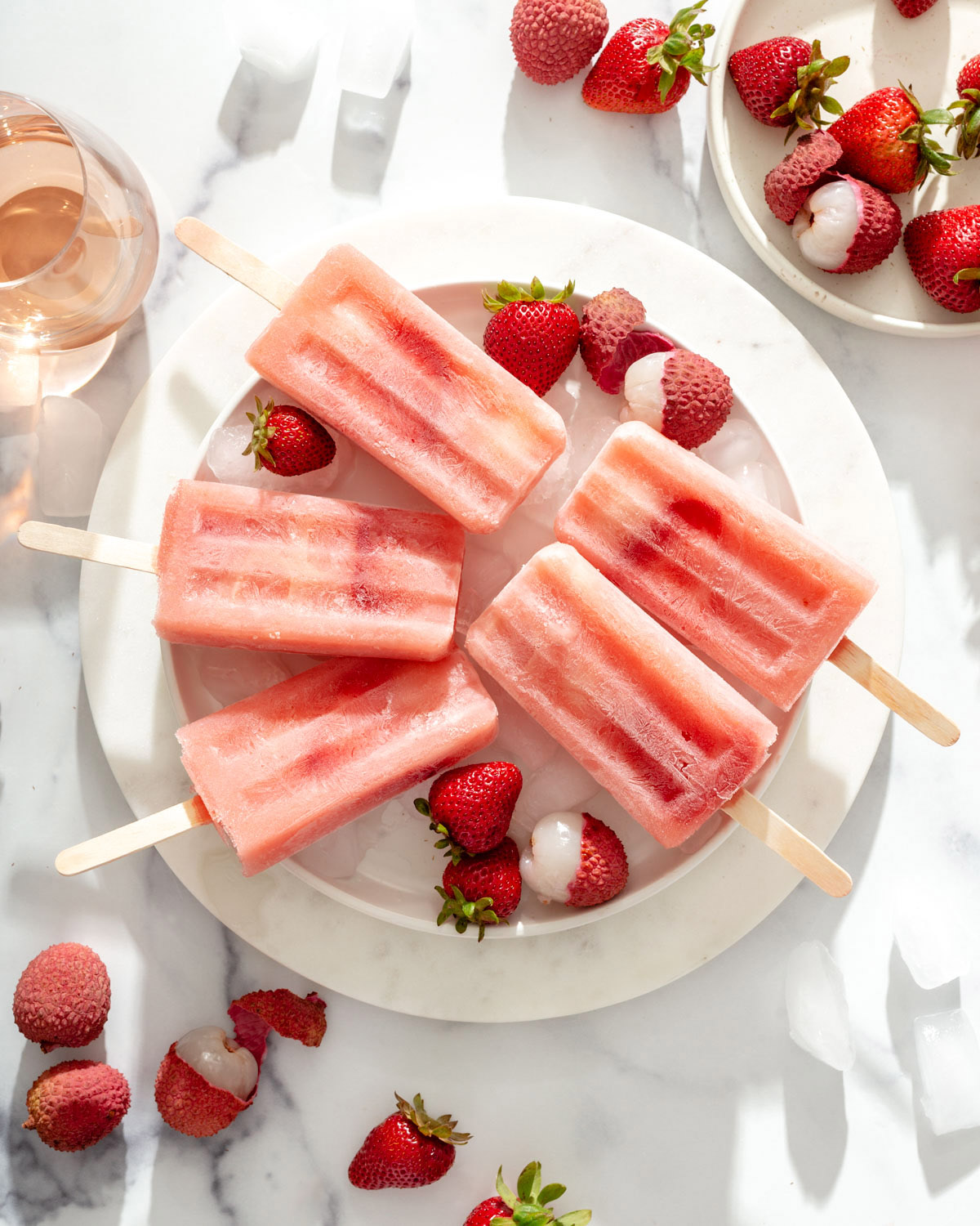 A plate of ice with popsicles resting on top of it with lychees and strawberries sprinkled around.