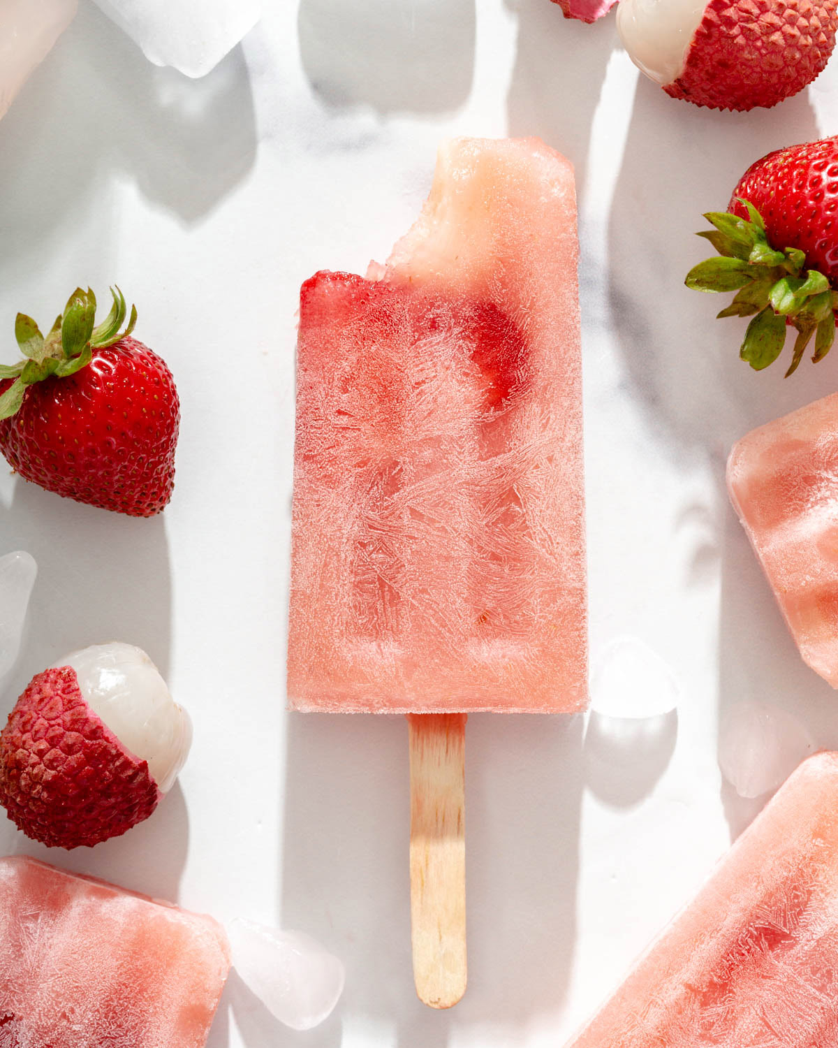 Close up of a bitten lychee frose popsicle on a table with popsicles and fruit around it.