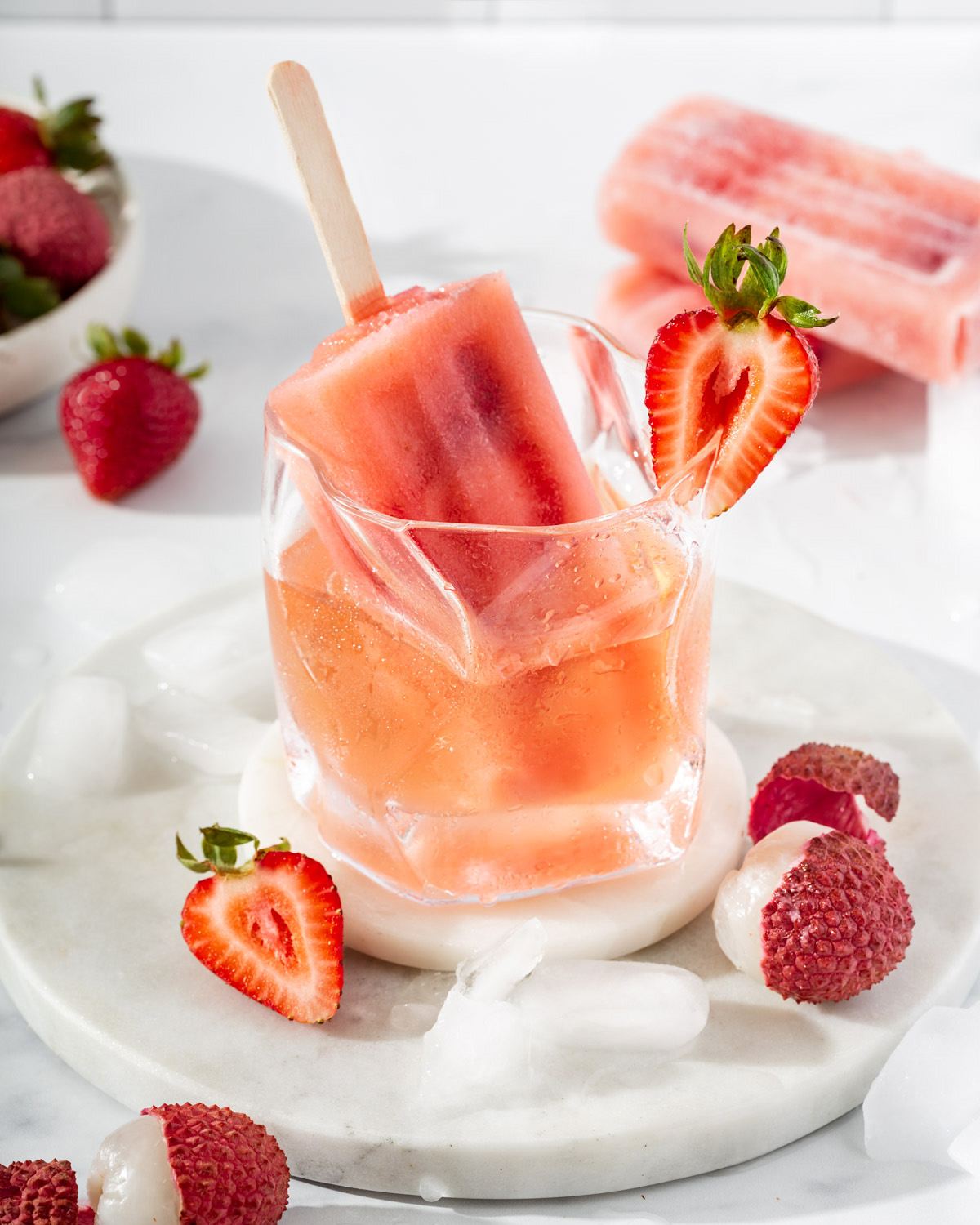 A frose popsicle sitting in a cocktail glass with rose surrounded by fruit and popsicles