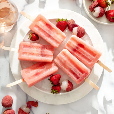 Close up of a plate of popsicles with fruit and a glass of rose around it.