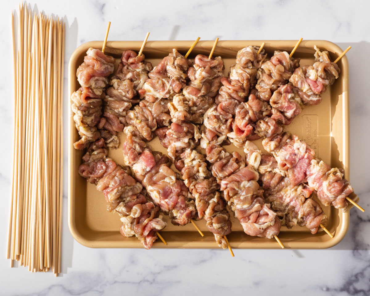 Marinated pork on skewers in a tray with skewers nearby.