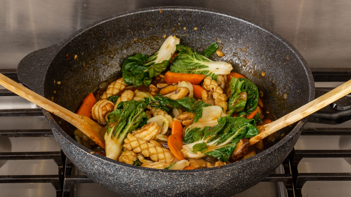 A wok full of proteins and vegetables being tossed with two wooden spatulas