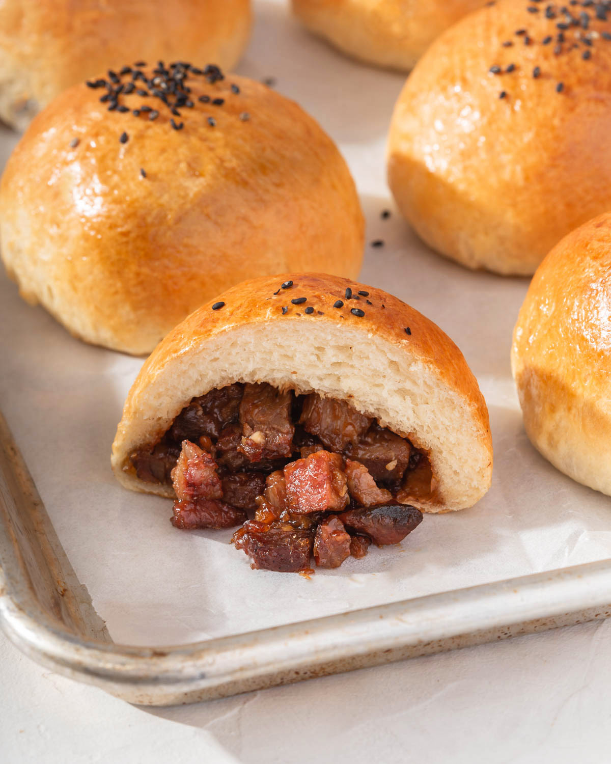 A sliced open BBQ pork bun sitting on a baking tray surrounded by more buns.