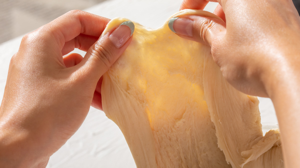 Someone pulling apart dough to show the flexibility of it with the "window pane test"