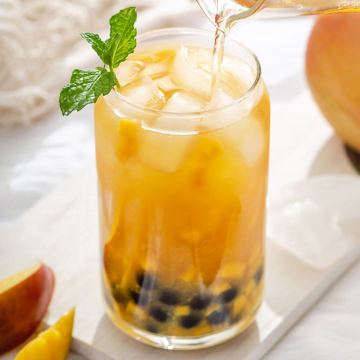 Up close of someone pouring green tea into a glass of mango green tea with boba and ice.