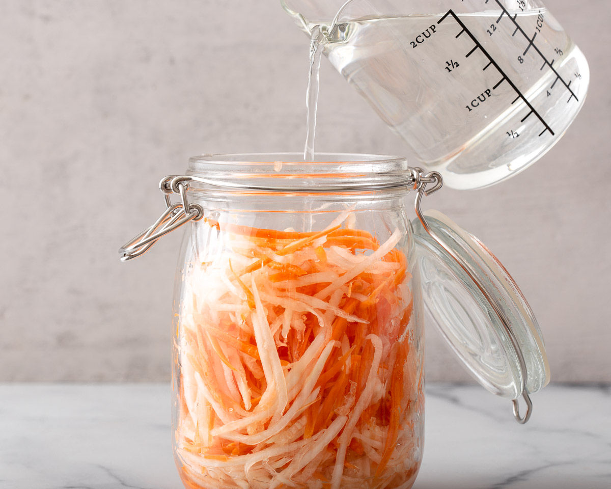 Pouring a sugar and vinegar solution into a jar of shredded carrots and daikon.