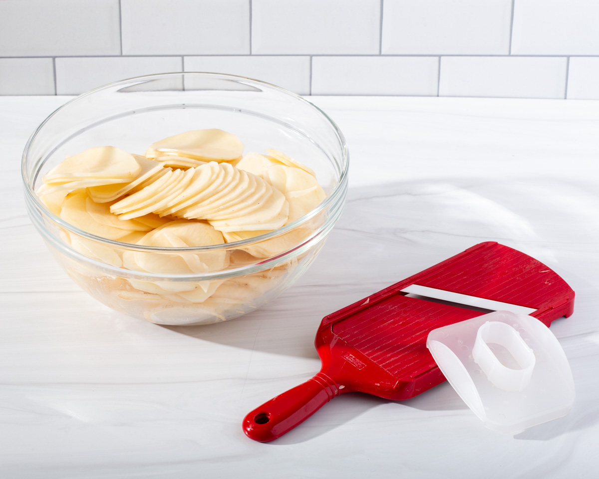 A mixing bowl full of thin sliced potatoes with a mandolin near by