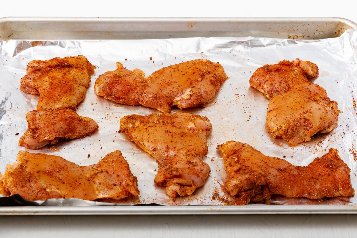 Chicken thighs throughly covered in seasoning on an aluminum foil lined tray