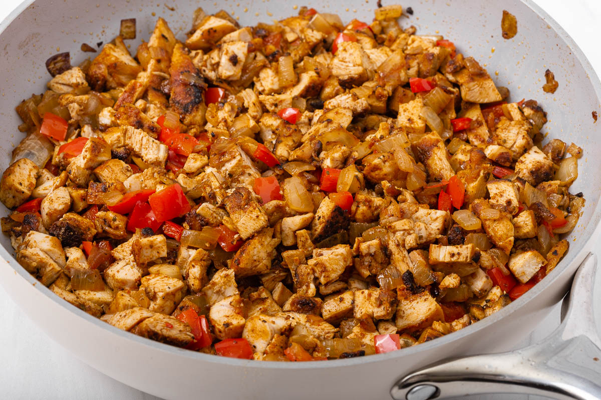 A skillet with the diced chicken tosses with caramelized onions and red bell peppers