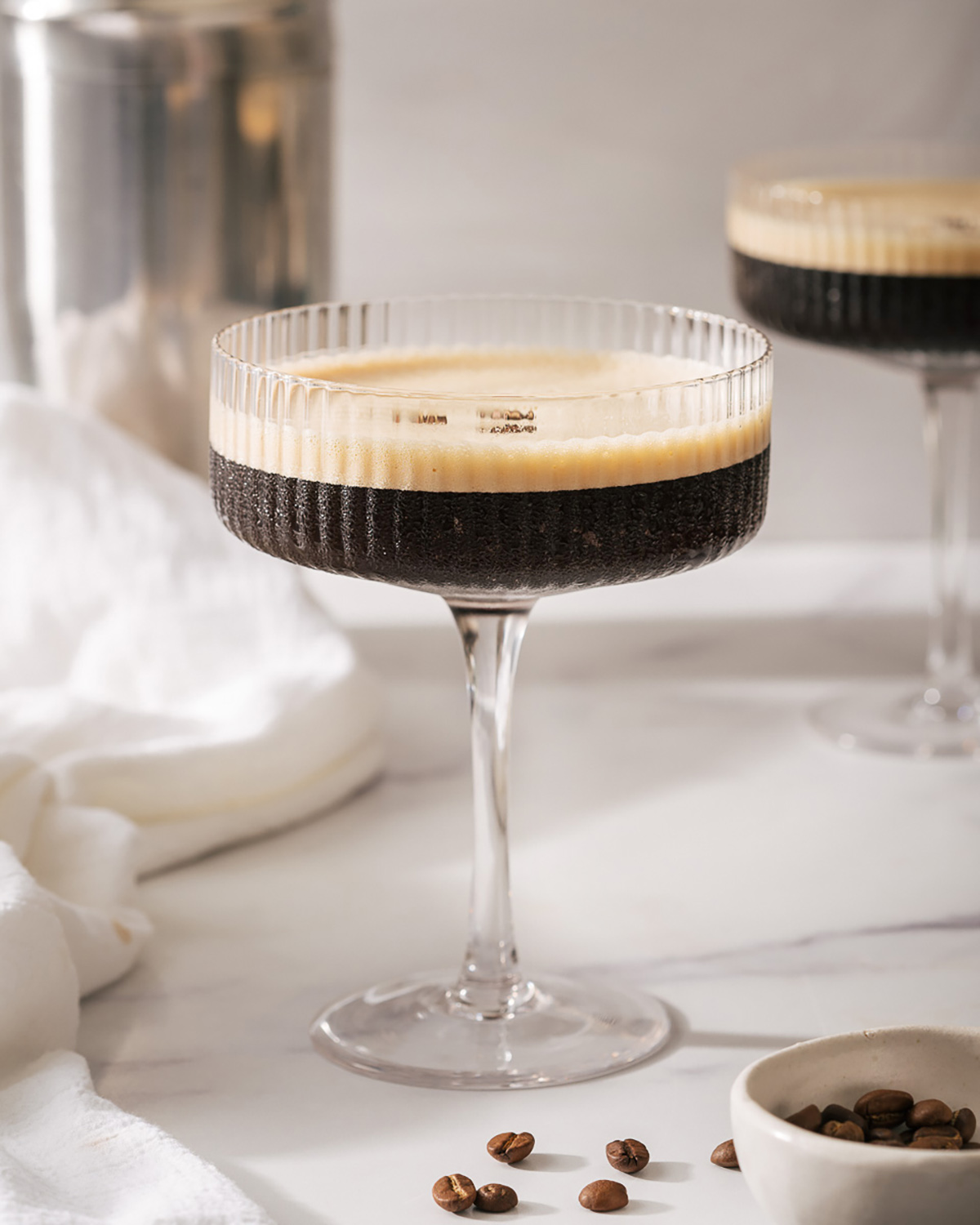 A martini glass with an espresso martini on a counter with another glass and shaker nearby
