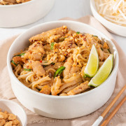 Up close of a bowl of chicken pad thai with sliced limes and chopsticks.