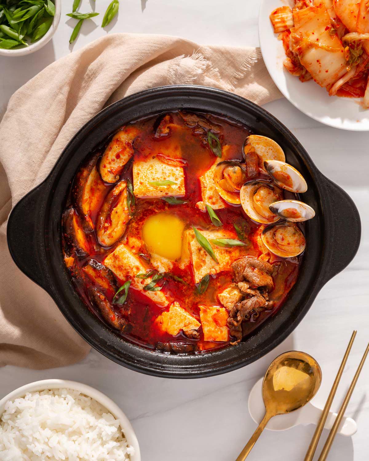 Looking down at a stone pot of spicy Korean tofu soup with kimchi and rice nearby.