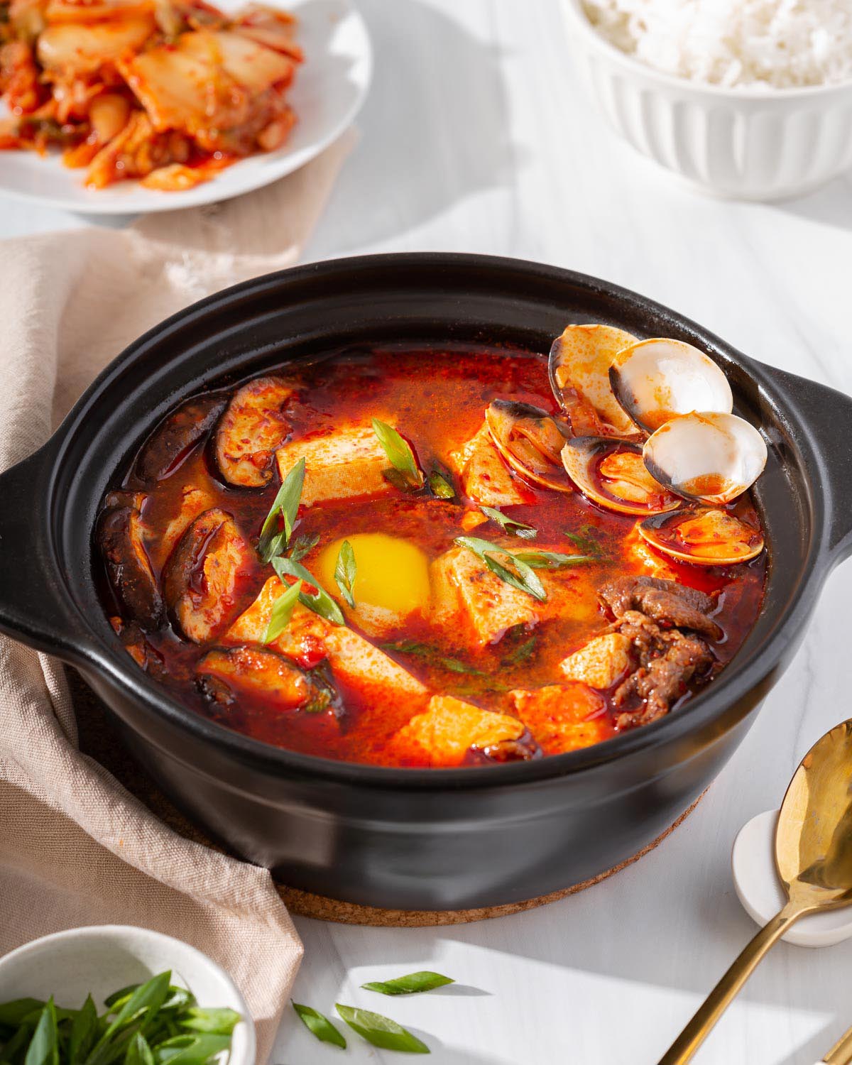 A stone pot of sundubu jjigae with kimchi, scallions, rice, and a spoon nearby.