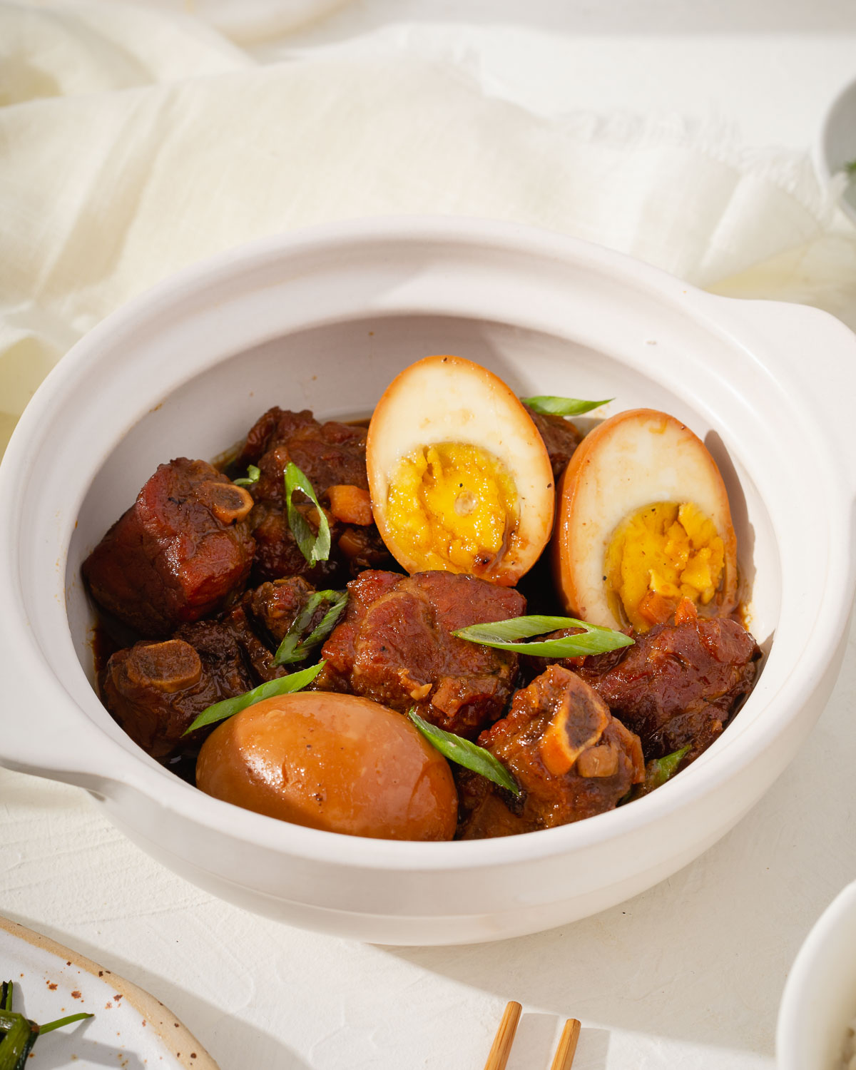 A white clay bowl of Vietnamese caramelized braised pork ribs with table settings nearby.