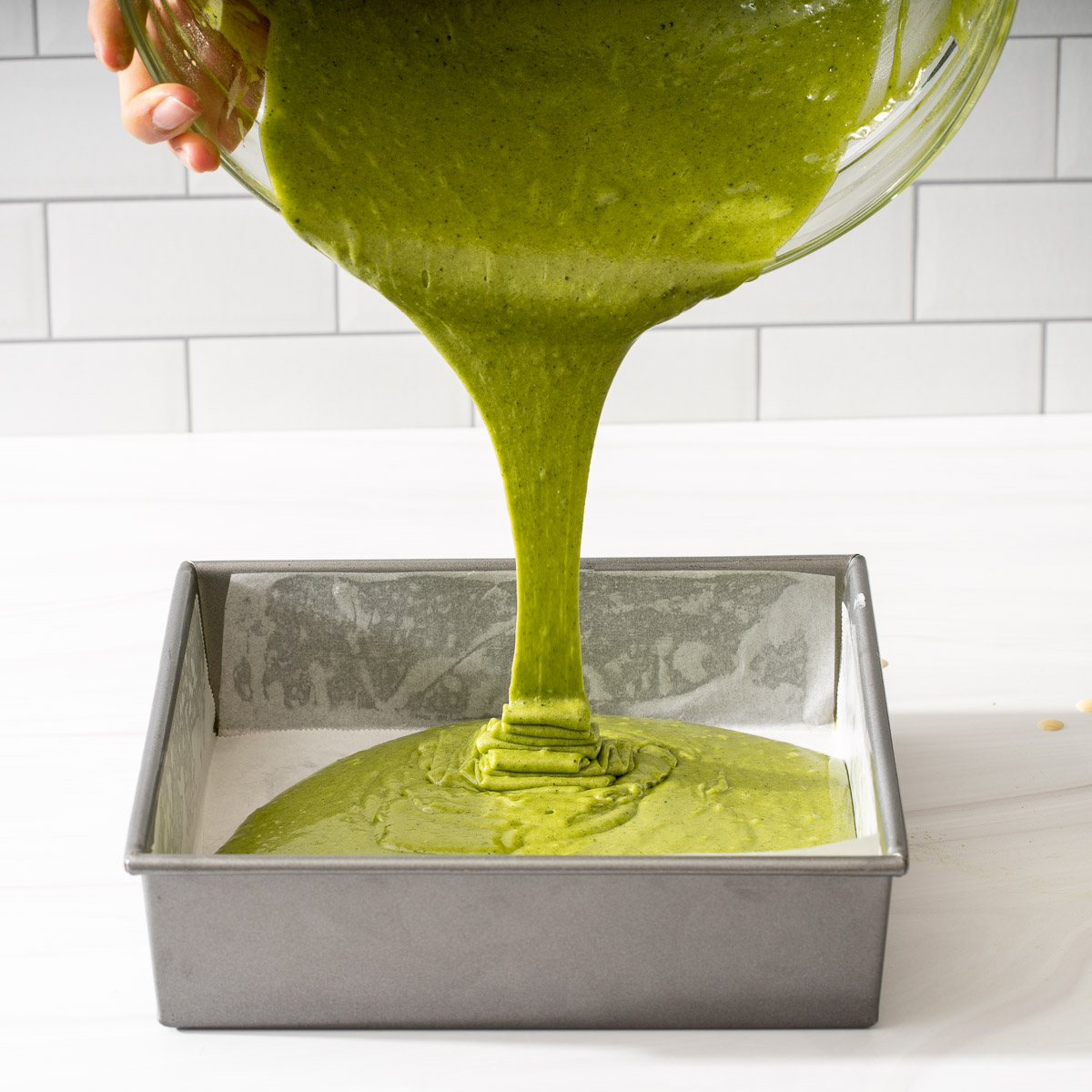 Pouring matcha brownie batter into a lined square baking pan.