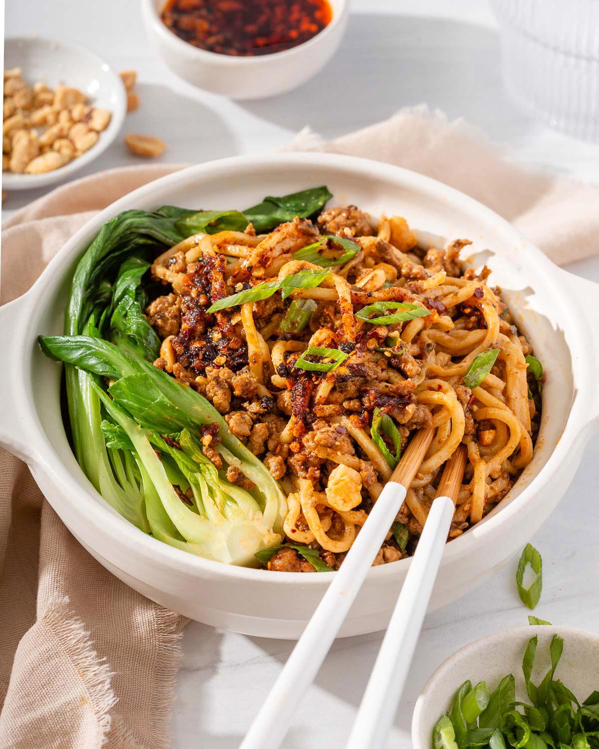Up close of a bowl of dan dan mian with peanuts, chili oil, and sliced green onions nearby.