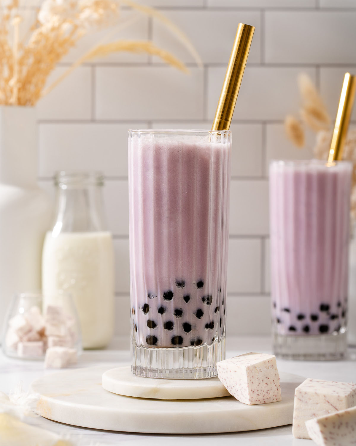 Two glasses of Taro milk tea on a counter with fresh taro root and milk nearby.