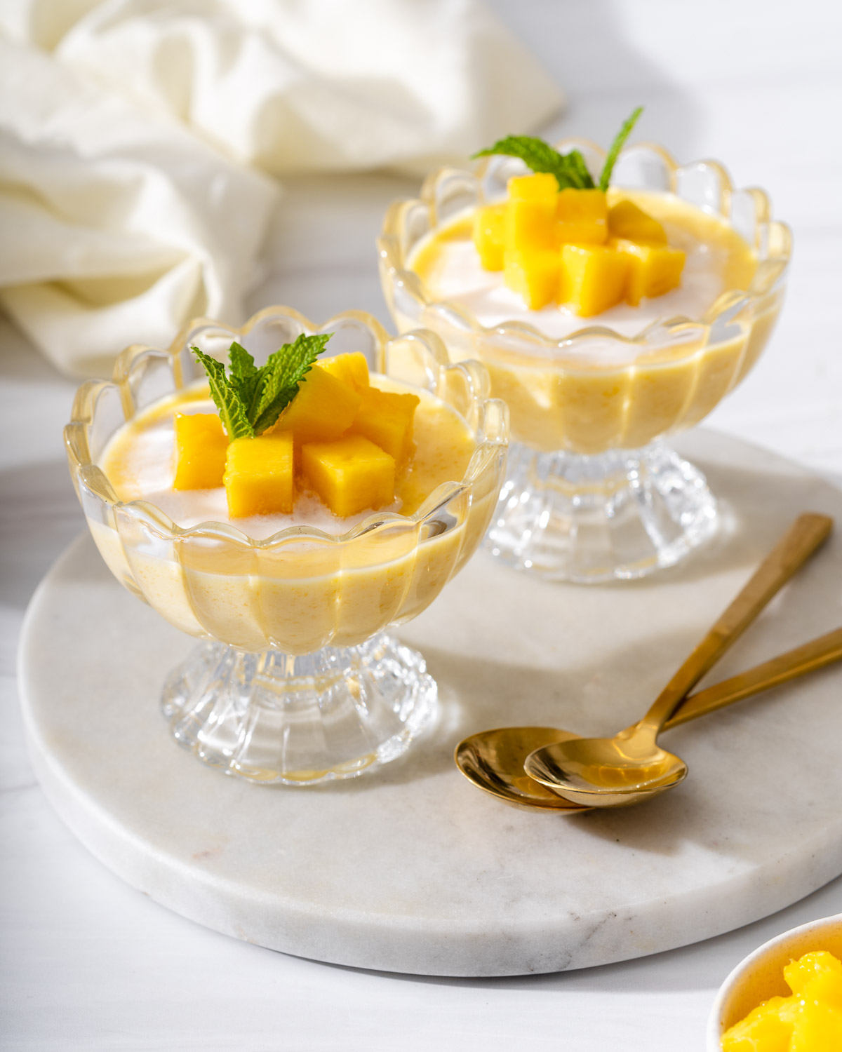 A serving tray with two dishes of mango pudding, fresh mango, and two spoons.