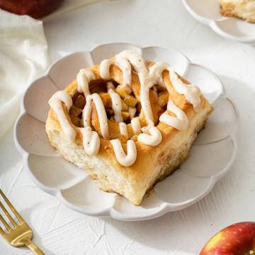 An apple cinnamon roll on a plate with utensils and apples nearby.