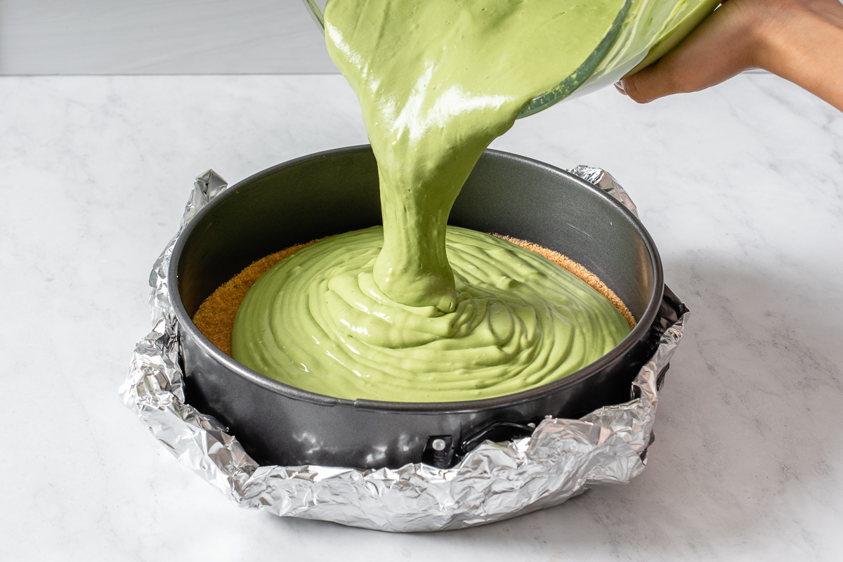 Pouring matcha cheesecake batter into a round baking dish with a graham cracker crust.