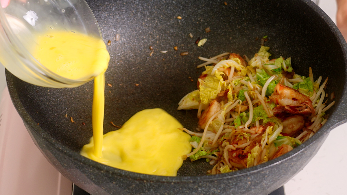 Pouring scrambled eggs into one half of a wok to make bihun goreng.