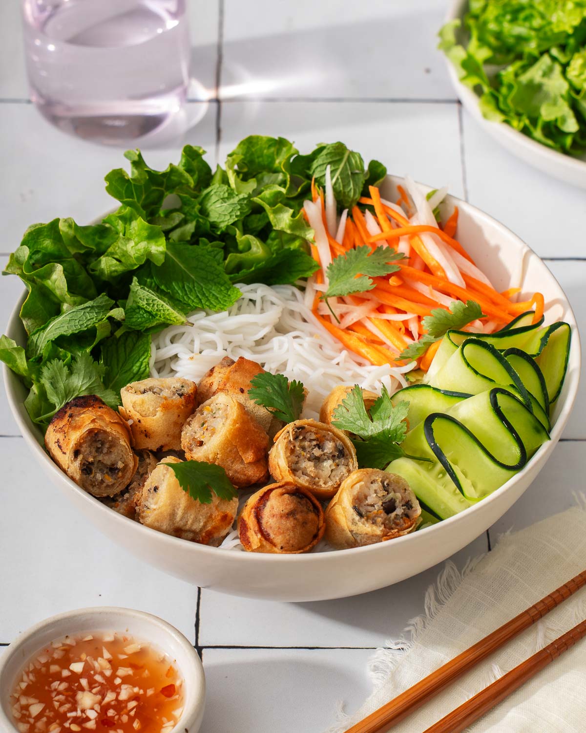 A bowl of vietnamese egg rolls with vermicelli noodles on a tile table.