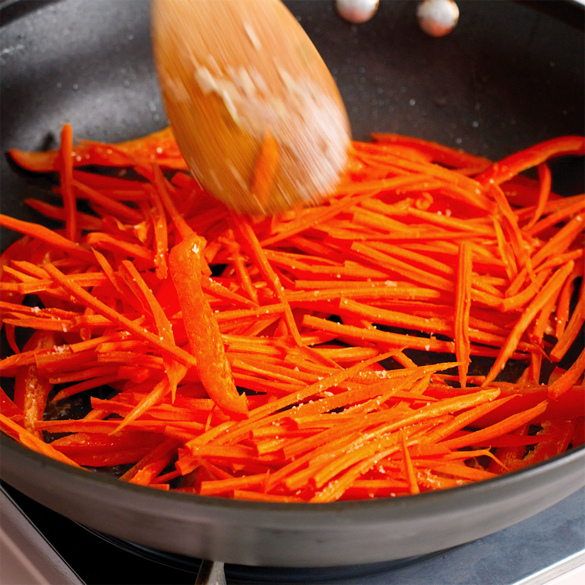 Cooking sliced red peppers and sliced carrots in a large skillet.