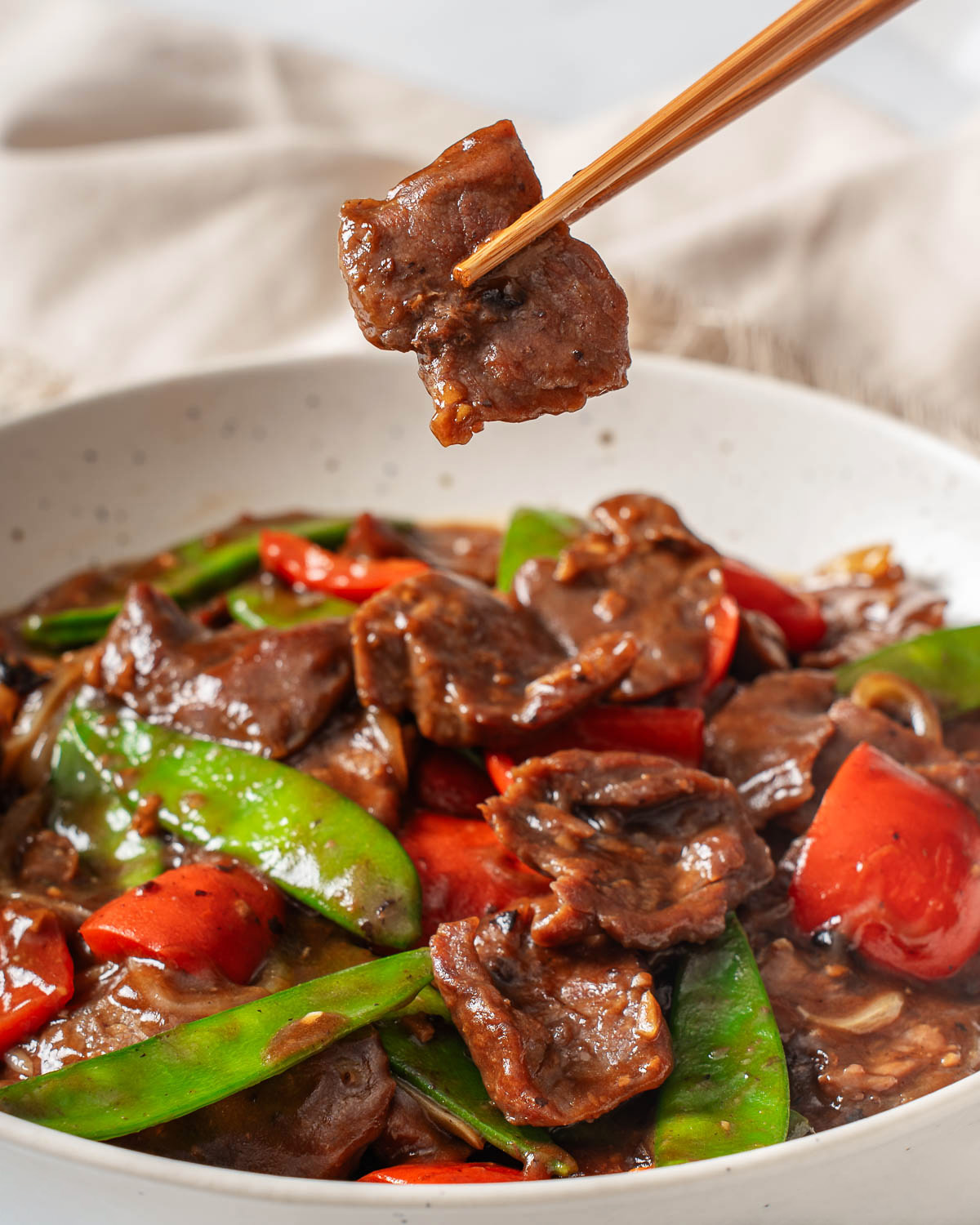 A pair of chopsticks lifting up a piece of steak from a plate of beef with black bean sauce.