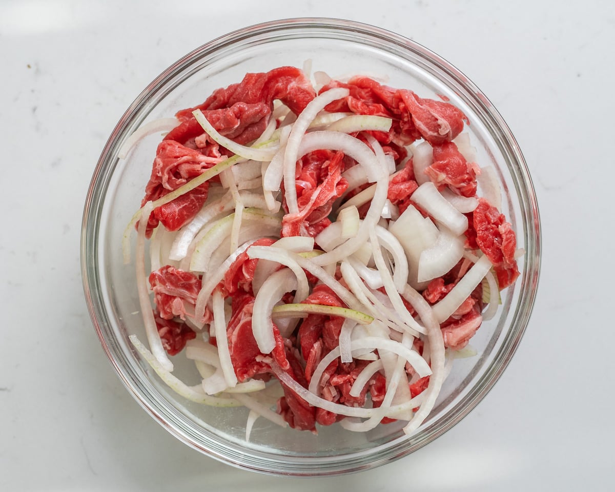 Thin sliced beef tosses with thin sliced yellow onion in a large glass bowl.