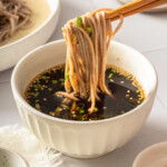Zaru soba being lifted from a bowl of tsuyu dipping sauce with chopsticks.