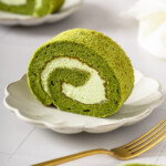 A matcha cake roll slice on a plate surrounded by table settings.