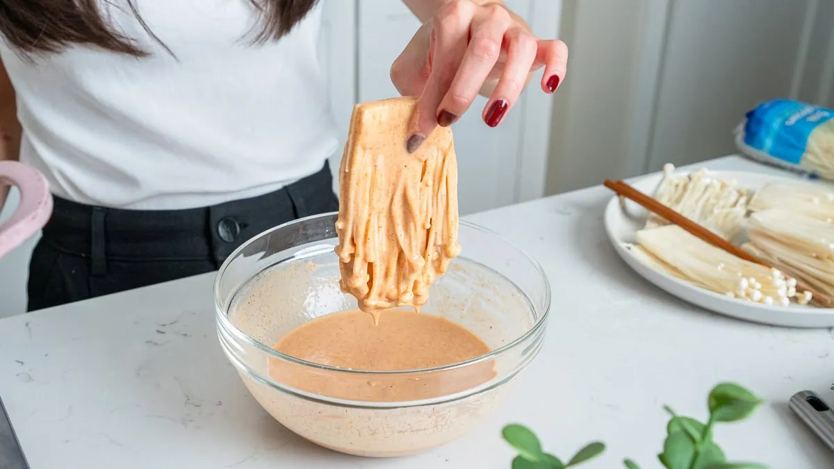 A slice of enoki mushroom being dredged in a wet batter.