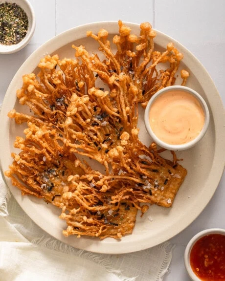 A top down view of a plate of fried enoki mushrooms with dipping sauces nearby.