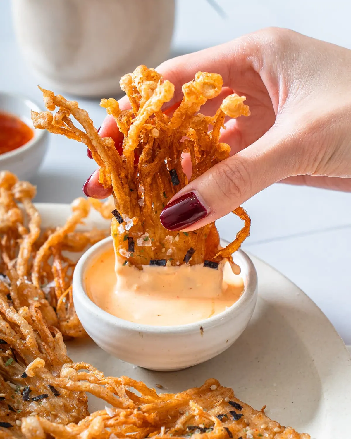 Someone dipping fried enoki mushrooms into a small dip bowl of sauce.