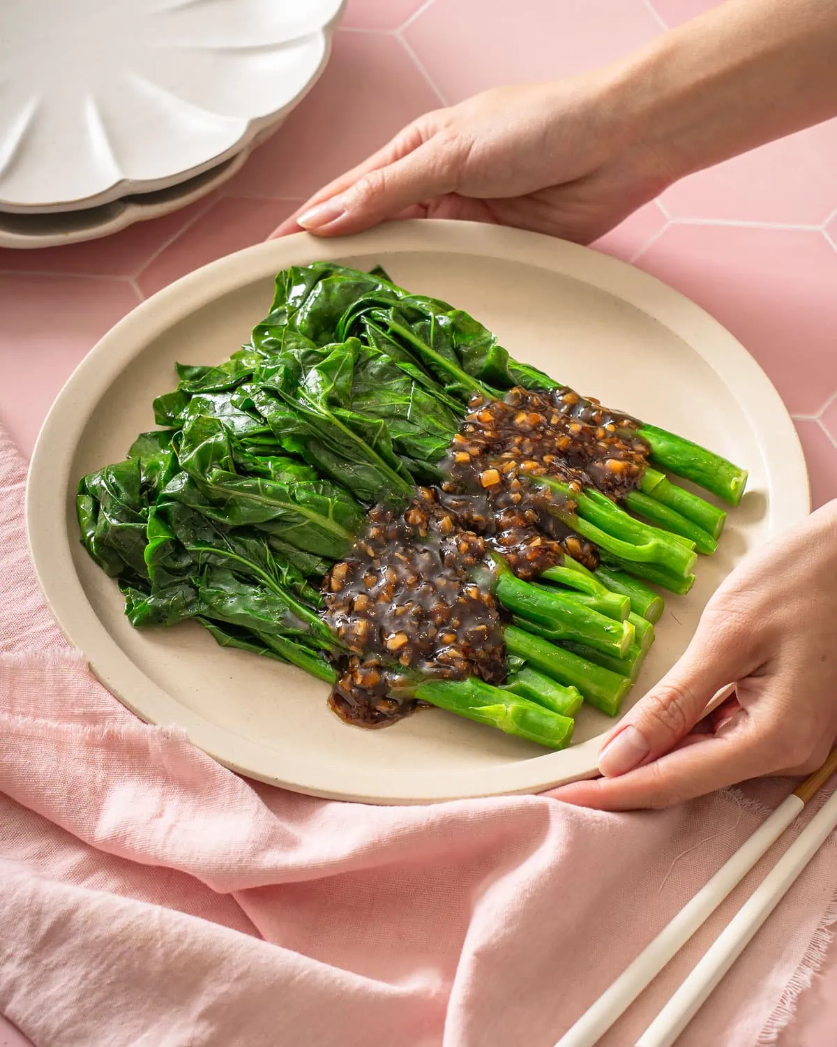 A pair of hands setting down a plate of gai lan with oyster sauce.