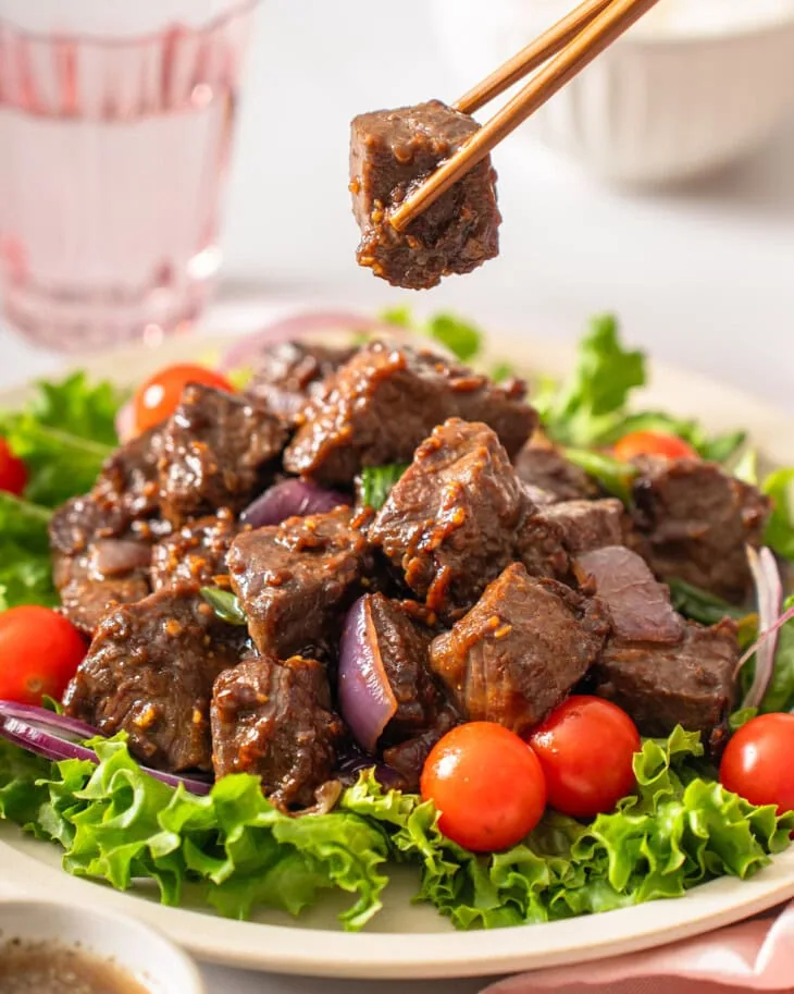 A pair of chopsticks lifting up a piece of Vietnamese shaking beef from a plate.