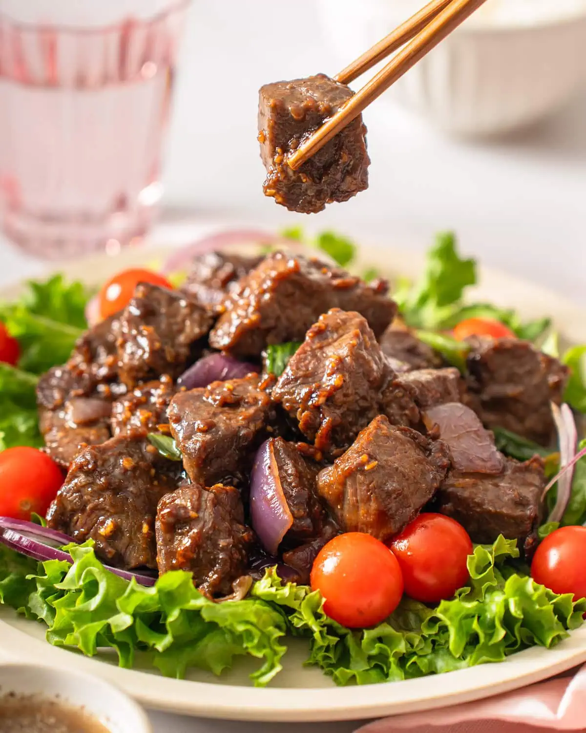 A pair of chopsticks lifting up a piece of Vietnamese shaking beef from a plate.
